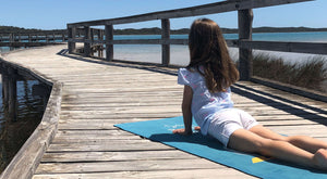 A young girl practicing yoga on the beach with a blue travel yoga mat on the peer in a cobra pose. This shows a yoga mat you can carry anywhere and where children can spend time on the mat to feel good.